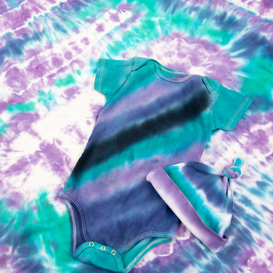 Teal, purple, and black tie dye baby set that includes a baby hat, bodysuit, and baby blanket.
