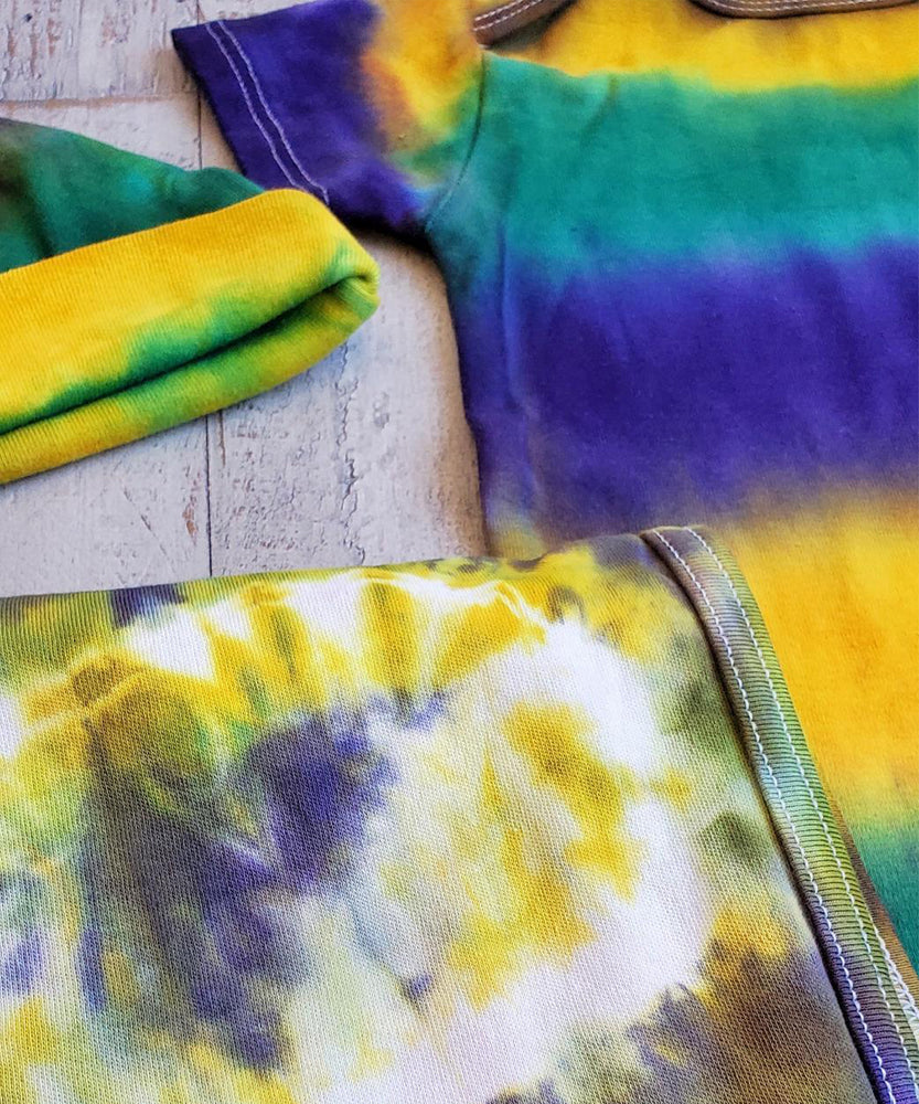 A tie dye organic baby set including a baby blanket bodysuit, hat, and blanket in Mardi Gras colors.