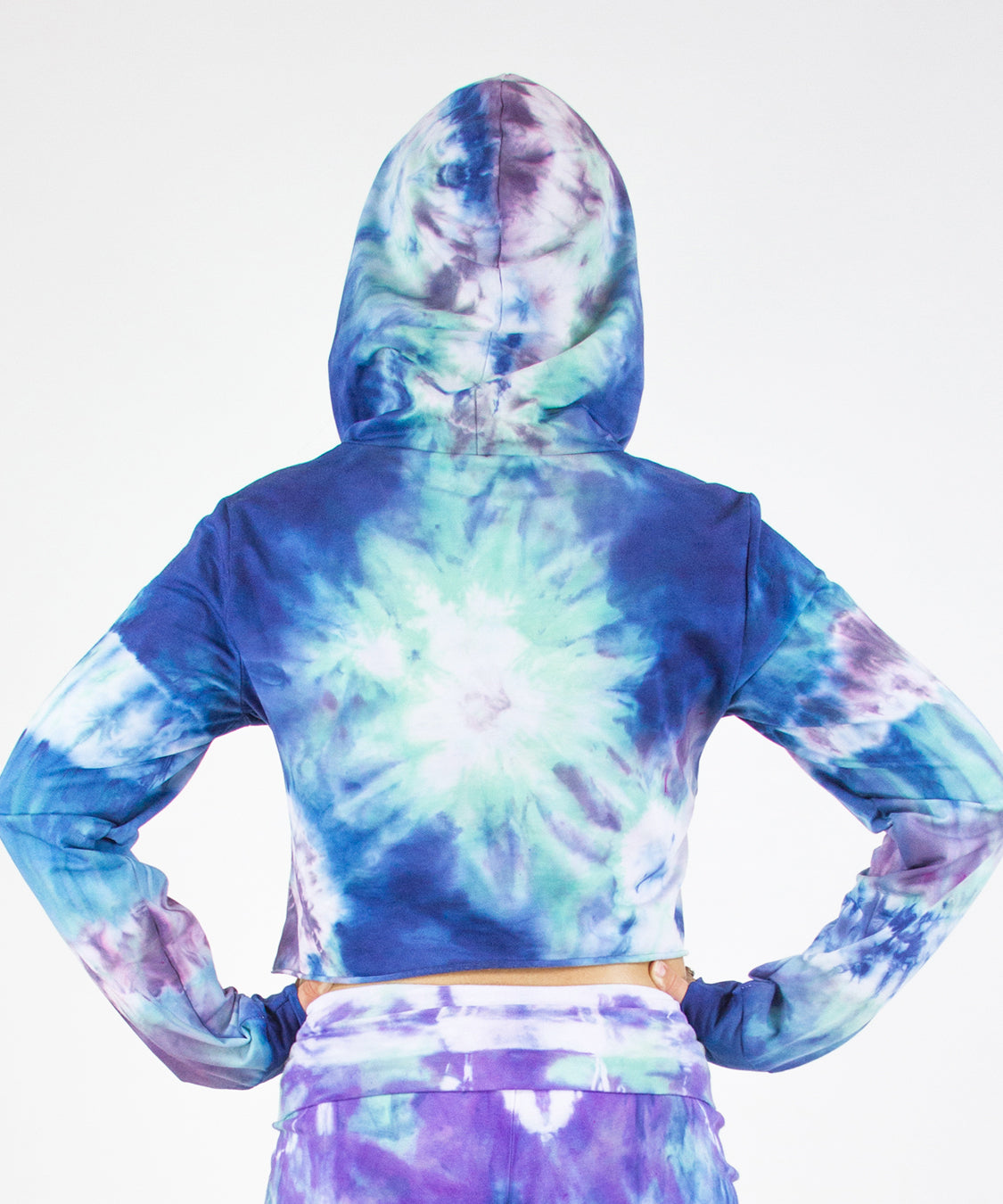 Woman wearing the Mykonos tie dye hoodie crop top featuring an oversized hood, drawstrings, and raw edge.  The colors in the crop top include blue, light teal, purple, and white.