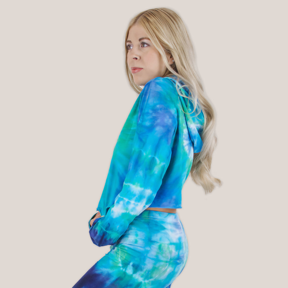 Woman wearing the Galapagos Islands Tie Dye Hoodie Crop Top.  The colors of the crop top include navy blue, teal, aqua, and blue.  Featuring a hood, drawstrings, and raw edge.