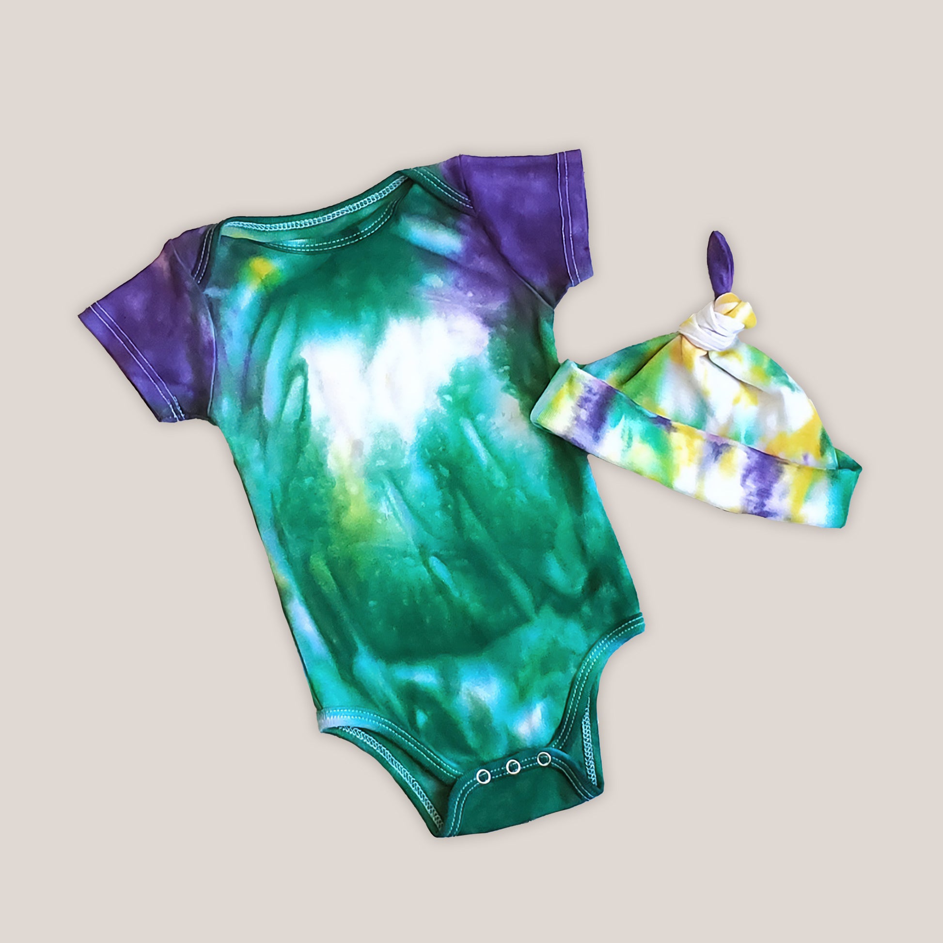 An organic tie dye baby bodysuit and hat in Mardi Gras colors of green, purple, and yellow.