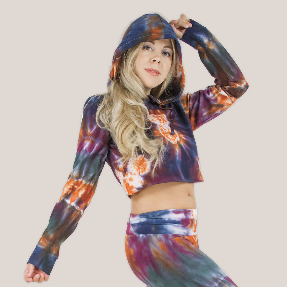 Woman wearing the Meteor tie dye hoodie crop top that features a hood, drawstrings, and raw edge.  The colors in the crop top include orange, navy, purple, and green by Akasha Sun