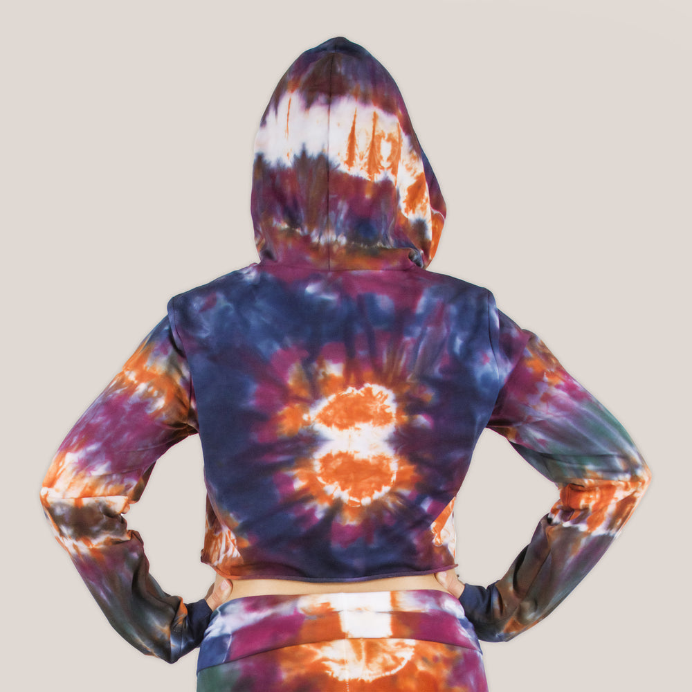 Woman wearing the Meteor tie dye hoodie crop top that features a hood, drawstrings, and raw edge.  The colors in the crop top include orange, navy, purple, and green by Akasha Sun