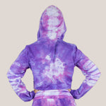 Woman wearing the Saint-Tropez tie dye hoodie crop top.  It features a hood, drawstrings, and a raw edge.  The colors in the top are purple, pink, and white.