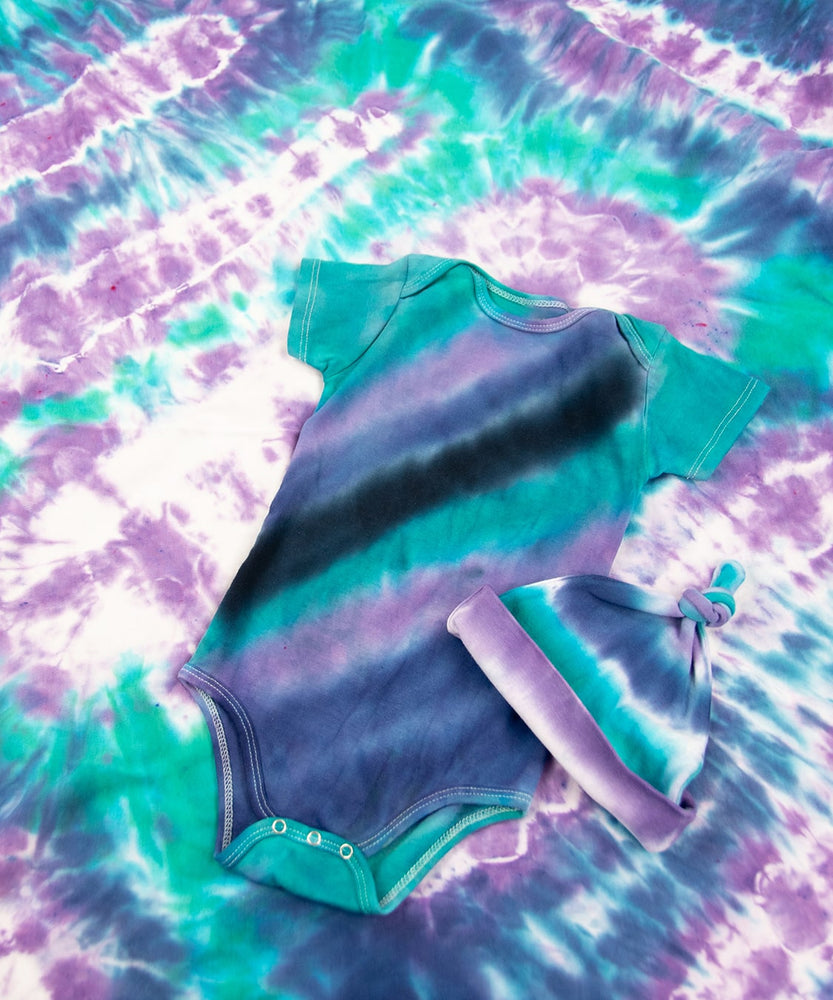 Teal, purple, and black tie dye baby set that includes a baby hat, bodysuit, and baby blanket.