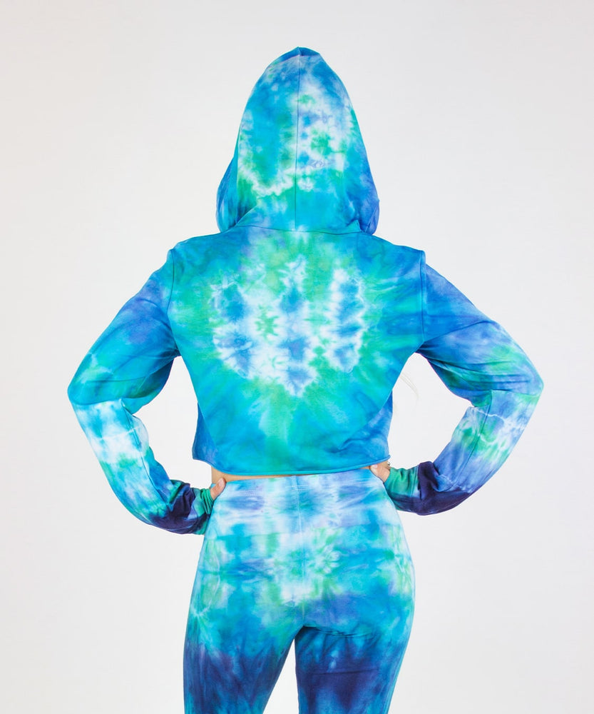 
                
                    Load image into Gallery viewer, Woman wearing the Galapagos Islands Tie Dye Hoodie Crop Top.  The colors of the crop top include navy blue, teal, aqua, and blue.  Featuring a hood, drawstrings, and raw edge.
                
            