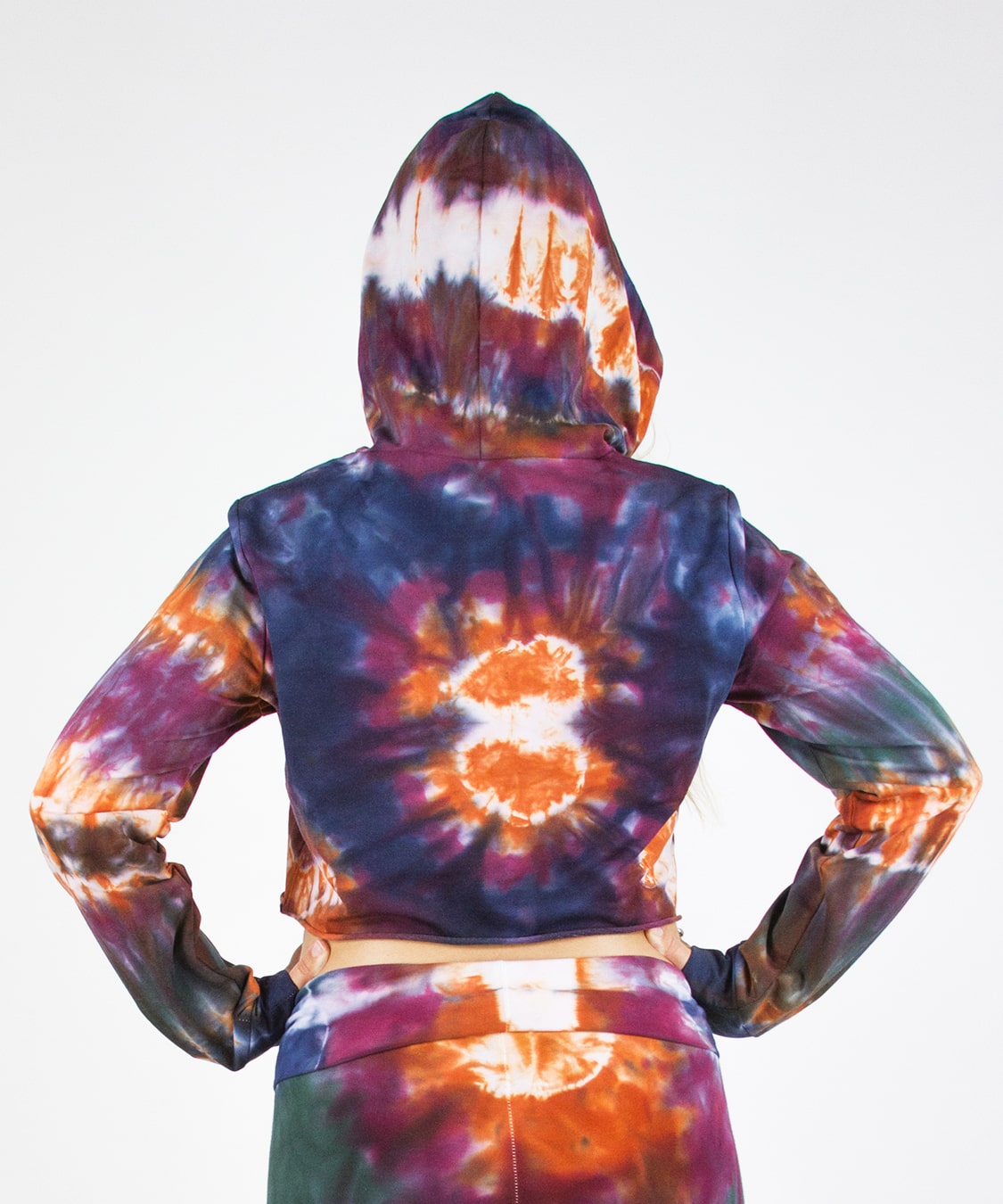 Woman wearing the Meteor tie dye hoodie crop top that features a hood, drawstrings, and raw edge.  The colors in the crop top include orange, navy, purple, and green.