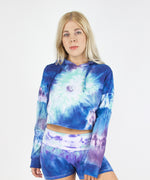 Woman wearing the Mykonos tie dye hoodie crop top featuring an oversized hood, drawstrings, and raw edge.  The colors in the crop top include blue, light teal, purple, and white.