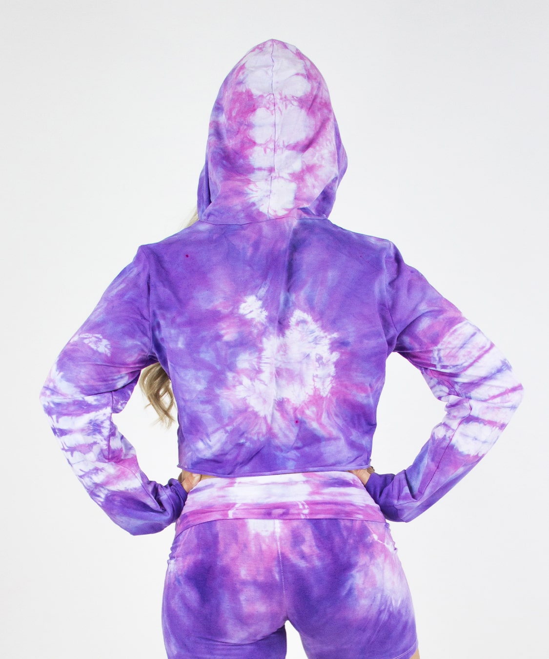 Woman wearing the Saint-Tropez tie dye hoodie crop top.  It features a hood, drawstrings, and a raw edge.  The colors in the top are purple, pink, and white.