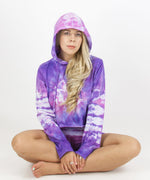 Woman wearing the Saint-Tropez tie dye hoodie crop top.  It features a hood, drawstrings, and a raw edge.  The colors in the top are purple, pink, and white.